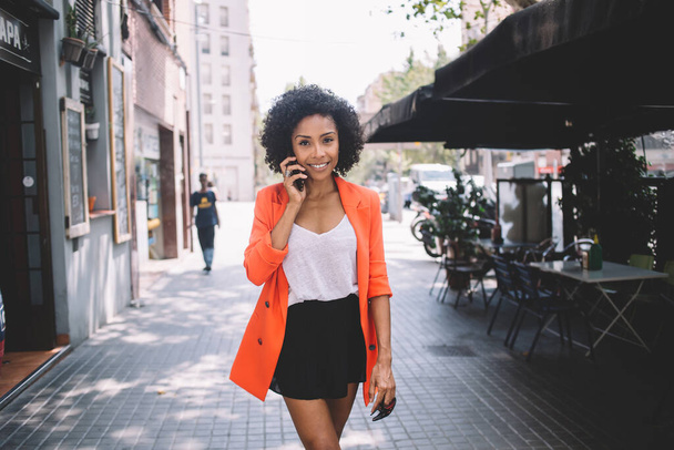 Happy young African American woman in bright summer clothes chatting on smartphone walking on city street on summer  sunny day - Fotó, kép