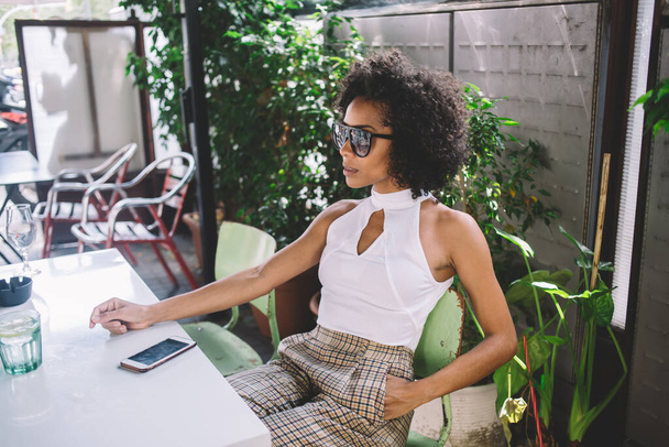 Jovem mulher afro-americana na moda em roupa da moda com óculos de sol e penteado afro sentado à mesa do café com smartphone olhando para longe da câmera - Foto, Imagem