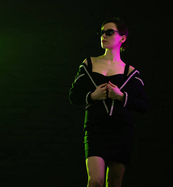 Young beautiful woman in fashionable sunglasses on a dark background in pink lighting. Place for text about fashion and beauty. - Fotó, kép