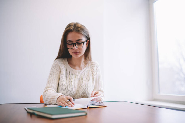 Young pensive female student in casual outfit with eyeglasses sitting at wooden table with book and flipping page while reading - Photo, Image