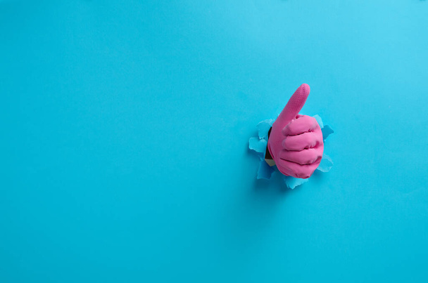 una mano in un guanto di gomma rosa per la casa sporge dal buco strappato e mostra un gesto ok, pollice in su. Fondo blu - Foto, immagini
