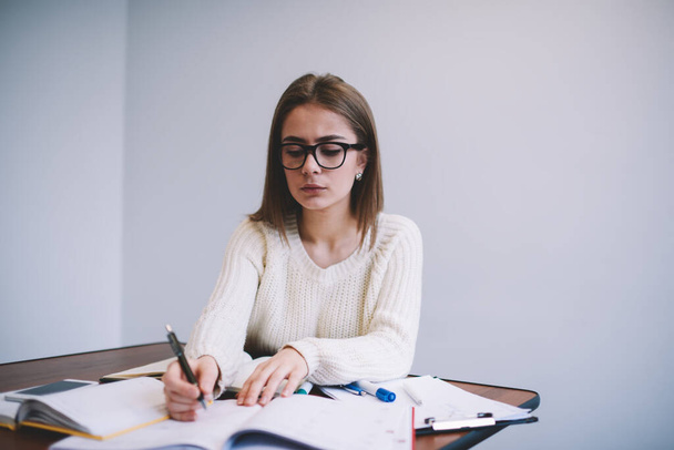 Focused female student in casual outfit with eyewear sitting at table and writing notes in copybook while doing task for university - Фото, изображение
