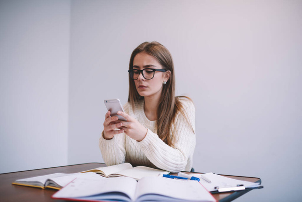 Concentrated young businesswoman in eyeglasses wearing white sweater text messaging on contemporary smartphone while working remotely with documents in light office - Fotoğraf, Görsel