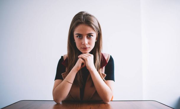 Wide angle of pensive female in casual clothes with long hair looking at camera while sitting at table with folded hands - Foto, immagini