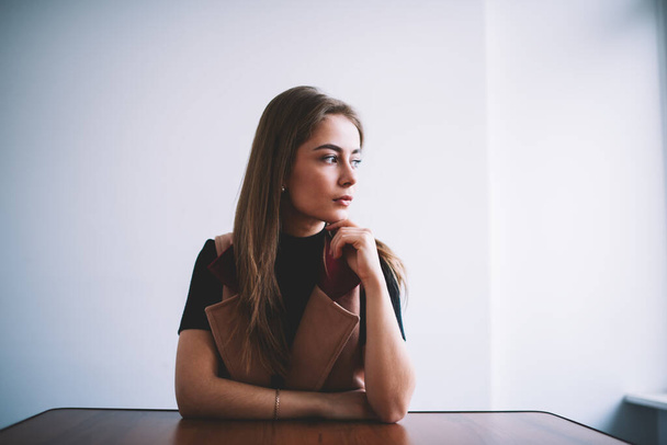 Thoughtful female in casual clothes looking away while sitting at table and touching chin in light room with white wall - Foto, Imagem