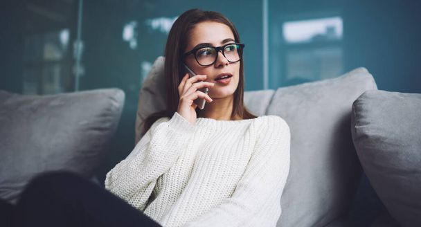 Young wistful female freelancer in casual sweater looking away while sitting on sofa and having conversation on smartphone during break - Zdjęcie, obraz