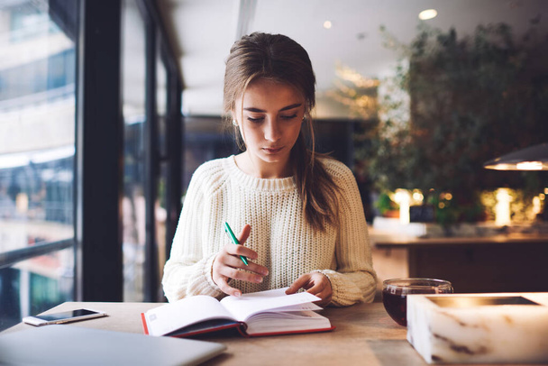 Pensive young woman in light sweater taking notes in notebook while sitting at table with cup of beverage near window in cafe in daytime - Foto, imagen
