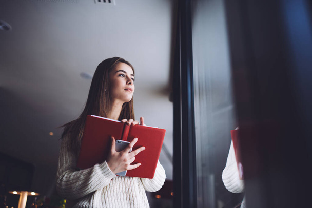 From below of thoughtful female entrepreneur in casual clothes standing with red notebook and smartphone and looking at window in daytime - 写真・画像