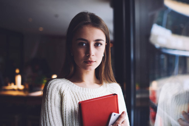 Young attentive female student in casual clothes with long hair looking at camera while standing with notepad and mobile phone - Fotoğraf, Görsel