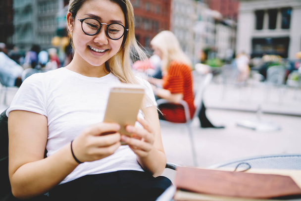 Happy Chinese blogger browsing website in social media spending leisure for smartphone networking, cheerful hipster girl in optical eyeglasses using mobile technology for text messaging and chatting - 写真・画像