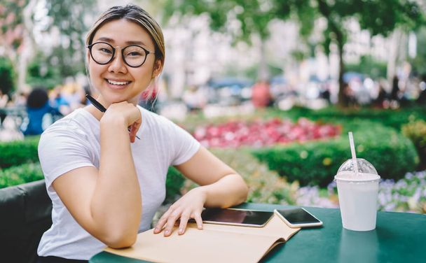Portrait of happy Asian student with education textbook for studying and takeaway beverage sitting at table and smiling at camera, cheerful hipster girl in optical spectacles posing in street cafe - Foto, imagen