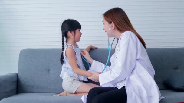 Азиатская красивая женщина-врач держит стетоскоп, чтобы проверить пульс, сердечный ритм милой маленькой девочки. Они оба сидели на диване. Концепция медицинских услуг в больницах, проверка здоровья детей. - Кадры, видео