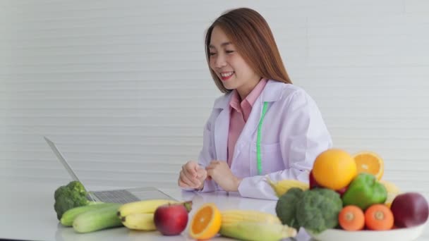 Gyönyörű ázsiai táplálkozási szakértő ül egy fehér íróasztal különböző gyümölcsök és zöldségek Ő ad ételt tanácsot a betegeknek. egy netes beszélgetésen keresztül egy laptopon. az egészségügyi ellátás fogalma - Felvétel, videó