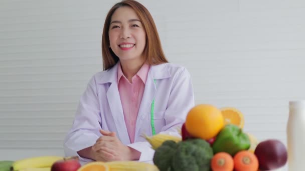 Жіночий лікар чи дієтолог, вродлива азіатка. Сидячи на білому столі з різними фруктами та овочами. Концепція охорони здоров'я, здорове харчування, втрата ваги - Кадри, відео