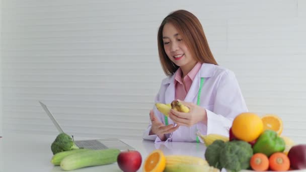 Gyönyörű ázsiai táplálkozási szakértő ül egy fehér íróasztal különböző gyümölcsök és zöldségek Ő ad ételt tanácsot a betegeknek. egy netes beszélgetésen keresztül egy laptopon. az egészségügyi ellátás fogalma - Felvétel, videó