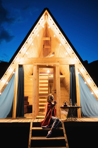 Vrouw rustend op de veranda van een houten zomer eco huis met lichten van bloemenslingers in de avond. Begrip glamping en verhuur van een chalet voor het weekend - Foto, afbeelding