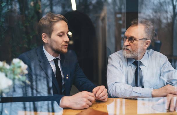 View through window of concentrated bearded mature male entrepreneur sitting with business partner wearing formal suit and discussing project details - Фото, зображення
