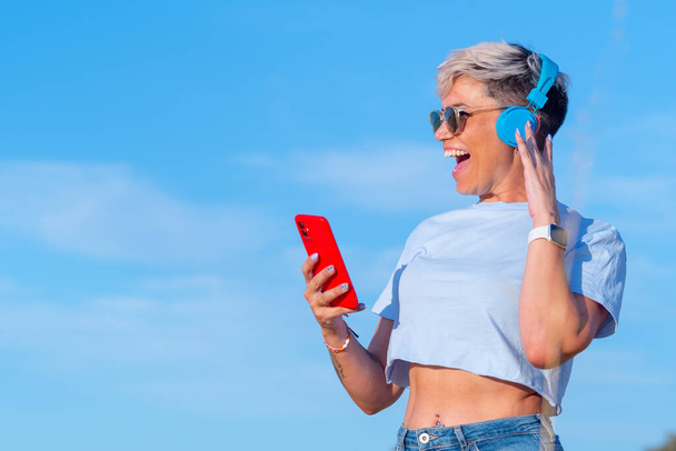 Lachende mooie jonge vrouw met kort blond haar gekleed in korte t-shirt tonen haar navel veel plezier tijdens het luisteren naar muziek met haar blauwe draadloze hoofdtelefoon en haar rode smartphone. - Foto, afbeelding