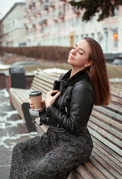 μια νεαρή γυναίκα με ένα μαύρο παλτό από δέρμα προβάτου με ένα φλιτζάνι καφέ μιας χρήσης στο χέρι της κάθεται σε ένα ξύλινο παγκάκι στο δρόμο χαμογελώντας. Το κορίτσι πίνει ένα ποτό. Καφές για την ιδέα - Φωτογραφία, εικόνα