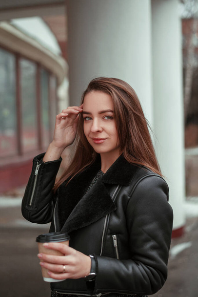 A young woman walks down an autumn winter city street and drinks takeaway coffee in a paper cup. The girl is drinking a drink. Coffee to go concept - Фото, зображення