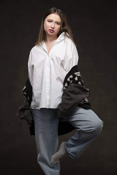 A beautiful girl in a white shirt and jeans stands on a gray background. - Foto, Imagen