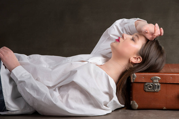 Красивая девушка лежит на старом чемодане на сером фоне. - Фото, изображение