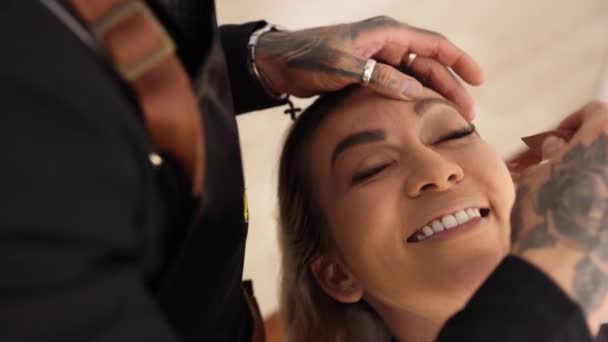 Van boven vrolijke vrouw glimlachen en tongzoenen tijdens make-up sessie met getatoeëerde visagiste in salon - Video