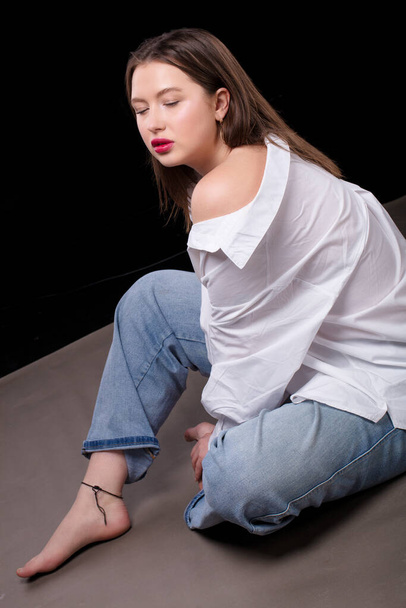 A beautiful girl in a white shirt and jeans sits on a gray background. - Foto, afbeelding