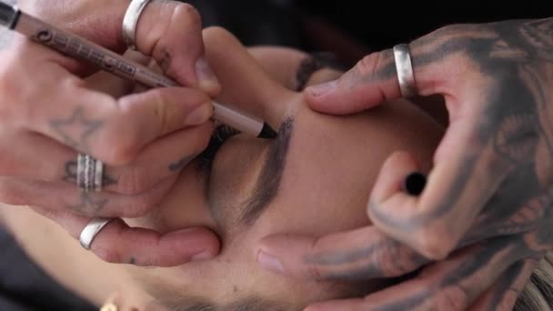 Dövmeli elleri olan yakın çekim makyaj sanatçısı kuafördeki kadın müşteriye mükemmel kaşlar çiziyor. - Video, Çekim
