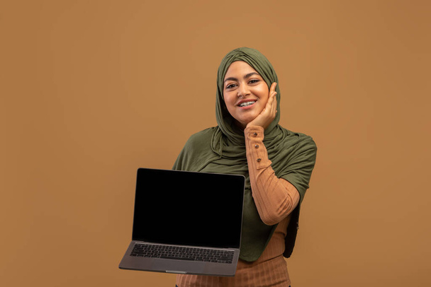 Nettitarjous. Iloinen arabi nainen osoittaa kannettavan tietokoneen tyhjä näyttö seisoo ruskea tausta, mockup - Valokuva, kuva