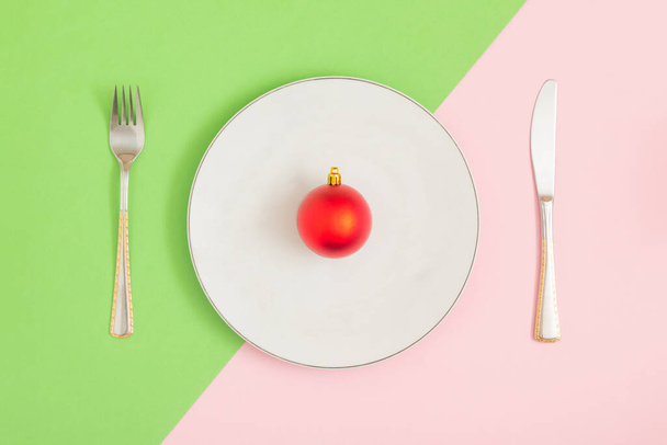 Ruokailuvälineet, siististi järjestetty värikkäälle pöydälle. Lautasella on punainen joulukoriste. Abstrakti luova koostumus. Uusi vuosi 2022 - Valokuva, kuva