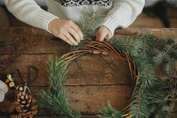 Φτιάχνω Χριστουγεννιάτικο στεφάνι. Γυναίκα σε άνετο πουλόβερ κρατώντας κλαδιά πεύκου και τακτοποίηση στεφάνι Χριστουγέννων σε ρουστίκ ξύλινο φόντο με κουκουνάρια, ψαλίδι. Προετοιμασία των χειμερινών διακοπών - Φωτογραφία, εικόνα