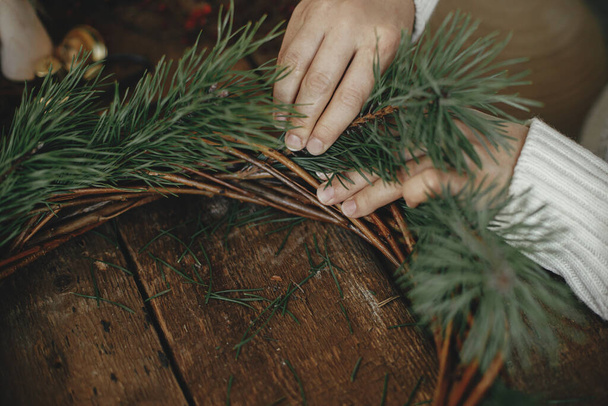 松の枝を持つ女性の手と素朴な木製の背景にクリスマスリースを配置します。冬休みの準備。お祭りのワークショップ。素朴なクリスマスの花輪を閉じる - 写真・画像
