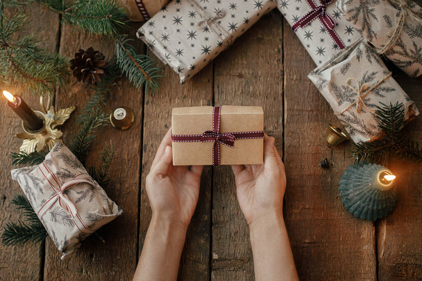 Hände halten ein stilvolles Weihnachtsgeschenk auf einem rustikalen Holztisch mit Kerzen, Scheren, Tannenzweigen. Stilvolle skandinavische Weihnachtsgeschenke, stimmungsvolles Stimmungsbild. Frohe Weihnachten und frohe Feiertage! - Foto, Bild