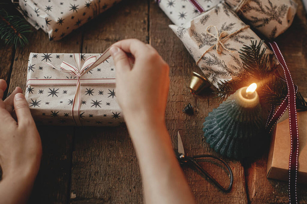 Hands opening stylish christmas gift on rustic wooden table with candles, scissors, fir branches. Merry Christmas! Stylish scandinavian xmas presents, atmospheric moody image - Photo, Image