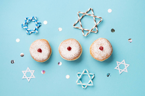 Ханука сладкие пончики sufganiyot с сахарной пудрой и фруктовым вареньем, праздничные звезды на голубом бумажном фоне. Еврейский праздник Ханука концепция. Вид сверху, пространство для копирования - Фото, изображение