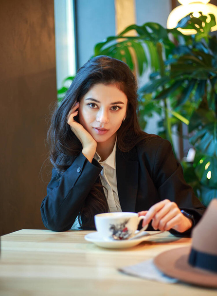 Young Asian business woman in a black jacket and hat in a cafe sits at a table with a cup of tea, on the background of a green plant - 写真・画像