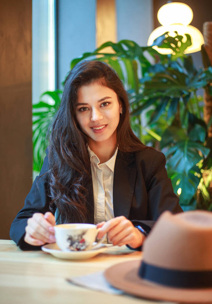 Young Asian business woman in a black jacket and hat in a cafe sits at a table with a cup of tea, on the background of a green plant - Zdjęcie, obraz