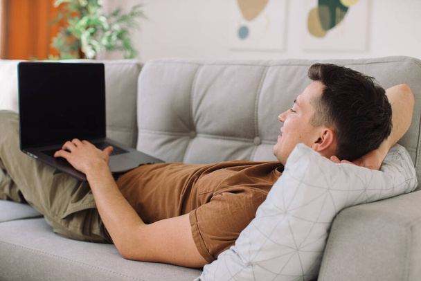 Красивый мужчина лежит на диване и работает над своим лаптоном - Фото, изображение
