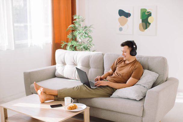 Kablosuz kulaklıklı rahat genç adam minimalist oturma odasında kanepede uzanıyor ve çevrimiçi çalışırken dizüstü bilgisayar kullanıyor. - Fotoğraf, Görsel