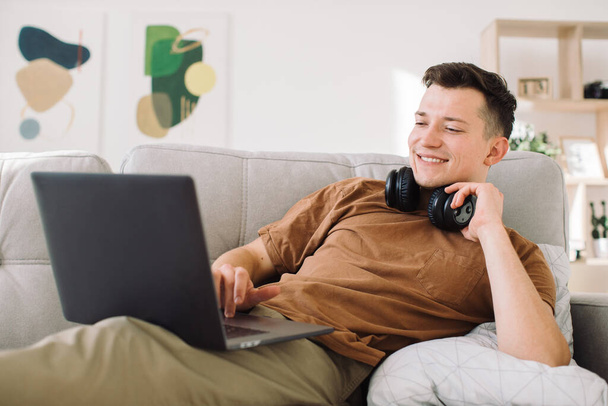 Χαλαρός νεαρός με ασύρματα ακουστικά ξαπλωμένος στον καναπέ στο μινιμαλιστικό σαλόνι και χρησιμοποιώντας φορητό υπολογιστή ενώ εργάζεται στο διαδίκτυο - Φωτογραφία, εικόνα