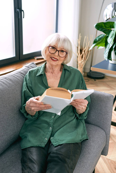 radosna starsza kobieta siedząca na kanapie i czytająca książkę w domu. Edukacja, dojrzała, koncepcja czasu wolnego - Zdjęcie, obraz