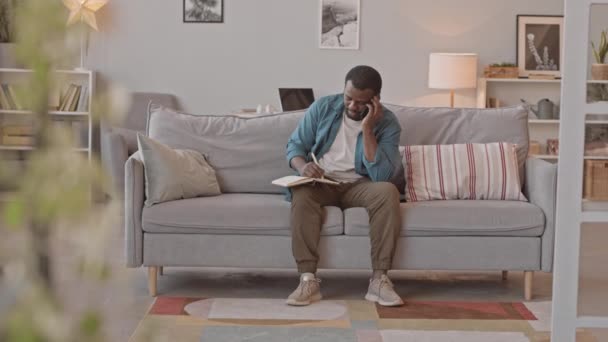 Slowmo shot di uomo afro-americano che parla su smartphone seduto sul divano in un accogliente soggiorno mentre le sue due piccole figlie giocose si divertono a giocare nelle vicinanze - Filmati, video