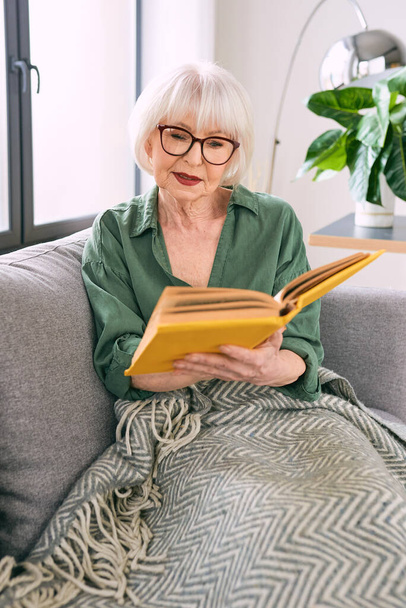 alegre anciana sentada en el sofá leyendo un libro en casa. Educación, madurez, concepto de ocio - Foto, Imagen