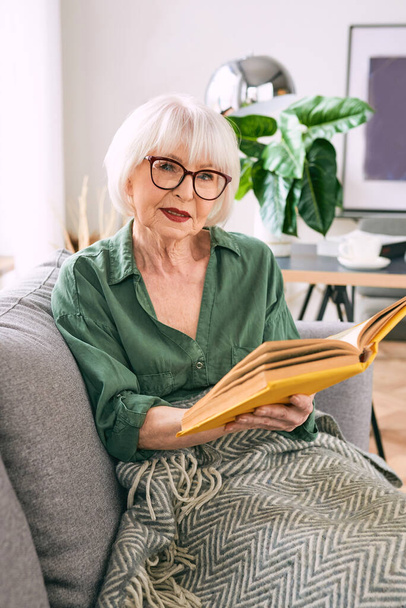 Χαρούμενη ηλικιωμένη γυναίκα που κάθεται στον καναπέ διαβάζοντας ένα βιβλίο στο σπίτι. Εκπαίδευση, ώριμη, έννοια αναψυχής - Φωτογραφία, εικόνα
