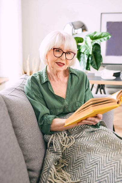 cheerful senior woman sitting on the couch reading a book at home. Education, mature, leisure concept - Photo, Image