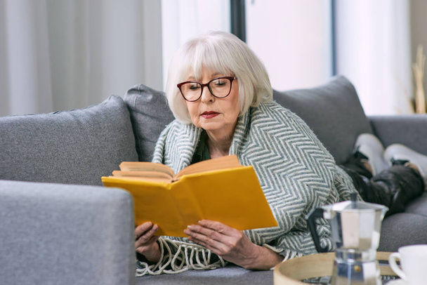 Egy régimódi idős nő fekszik a kanapén, és könyvet olvas. Oktatás, érett, szabadidős koncepció - Fotó, kép