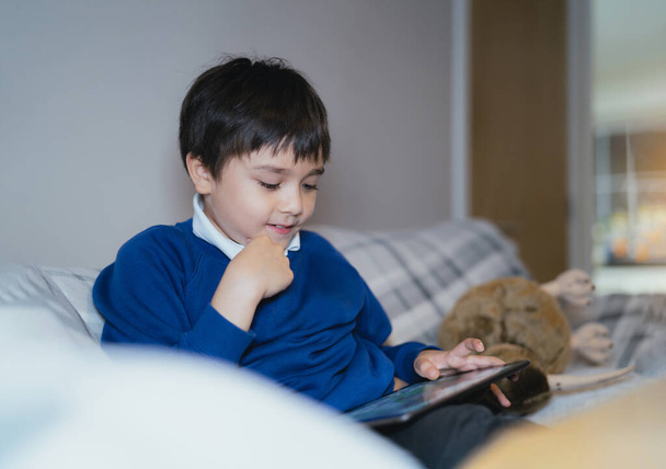 Happy schoolboy with smiling face playing game online with friend on tablet,Kid using internet sending homework to the teacher,Positive child sitting on sofa relaxing alone in living room after School - Foto, immagini