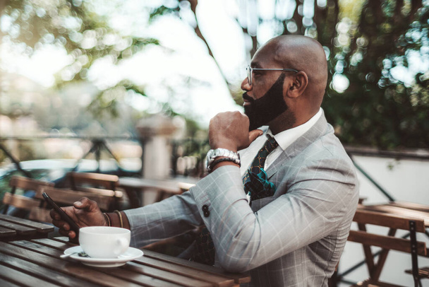 A fashionable mature bald African American businessman with a black well-groomed beard, in an elegant checkered costume, with a smartphone in his hand is having a coffee break in an outdoor restaurant - Photo, image