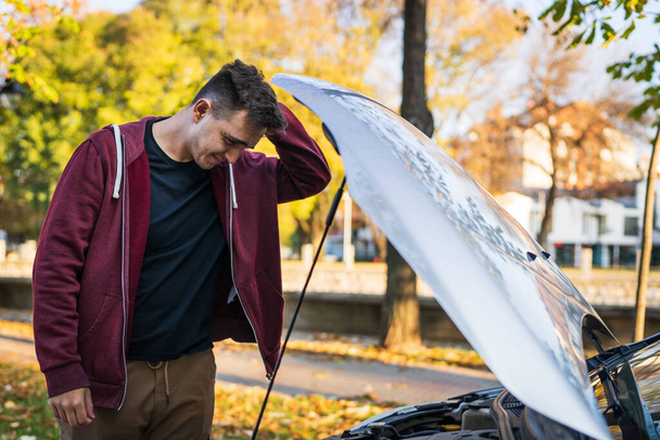 one Adult caucasian man standing by his car checking failed engine of broken vehicle holding his head in autumn day side view copy space - Фото, изображение